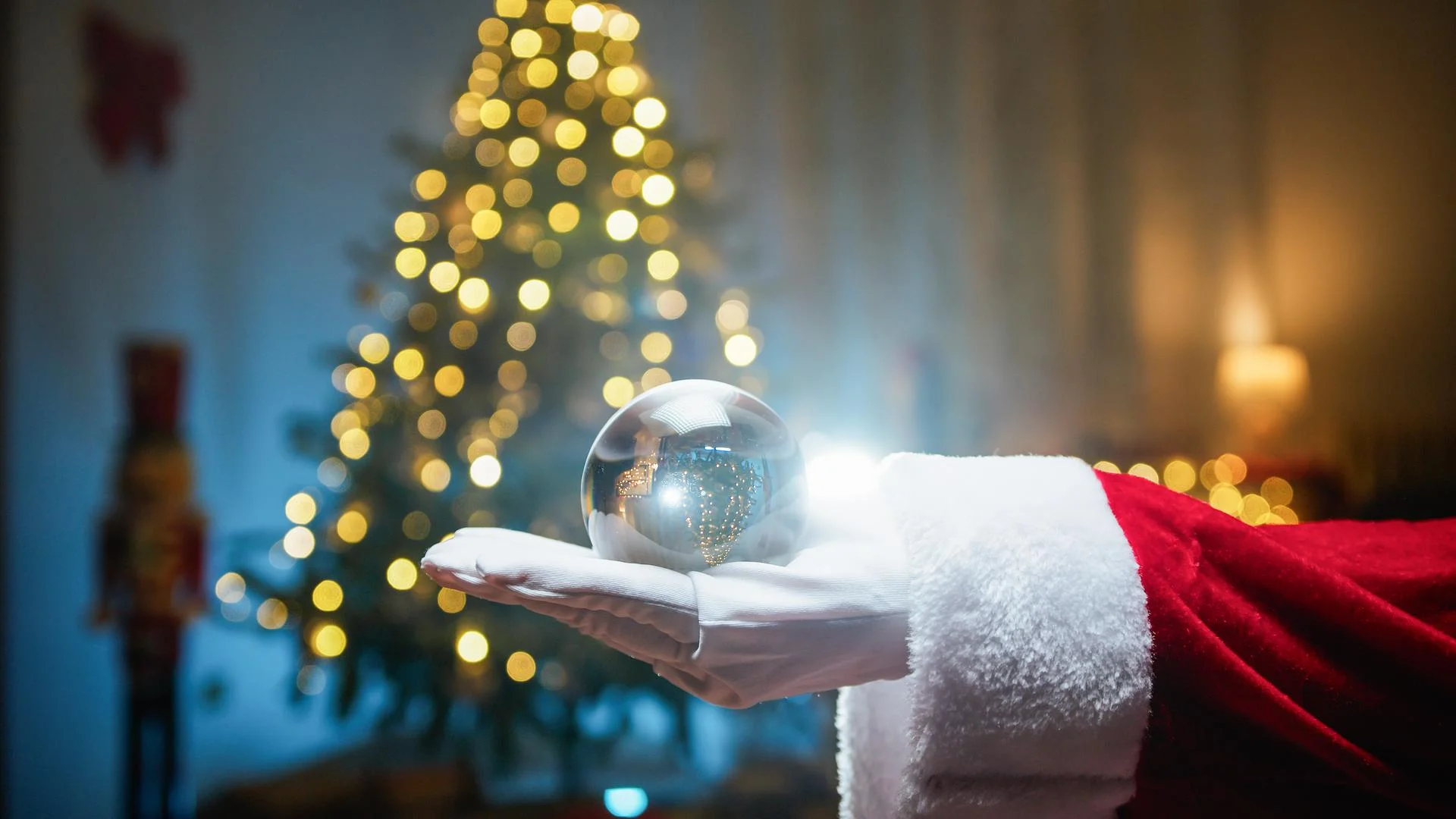 Santa’s gloved hand holding a sparkling glass ornament beside the warm lights of a Christmas tree.